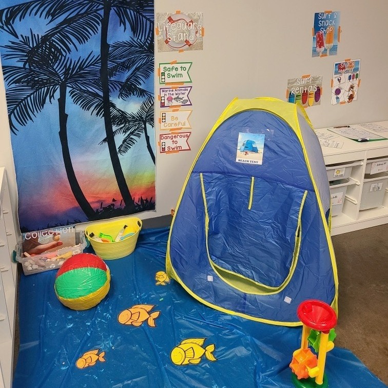 A corner of a room turned into a beach scene with a palm tree backdrop, pretend water and fish on the floor and a beach tent.